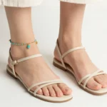 A woman's ankles adorned with a stacked set of gold chain and beaded turquoise anklets, paired with strappy beige sandals, showcasing a modern bohemian jewelry styling idea for summer outfits.