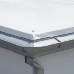 Close-up of a flat roof with a white TPO membrane installed, showing the proper transition and flashing where the roof edge meets a grey aluminum gutter system for effective drainage.
