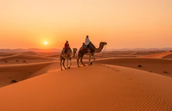 A Morocco desert tour camel trek at sunset on the orange sand dunes of Erg Chebbi near Merzouga, featuring a Berber guide and a desert camp in the Sahara.