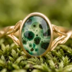 Nature-inspired moss agate engagement ring with green inclusions set in a yellow gold branch and leaf band, resting on moss and ferns for a spring botanical concept.