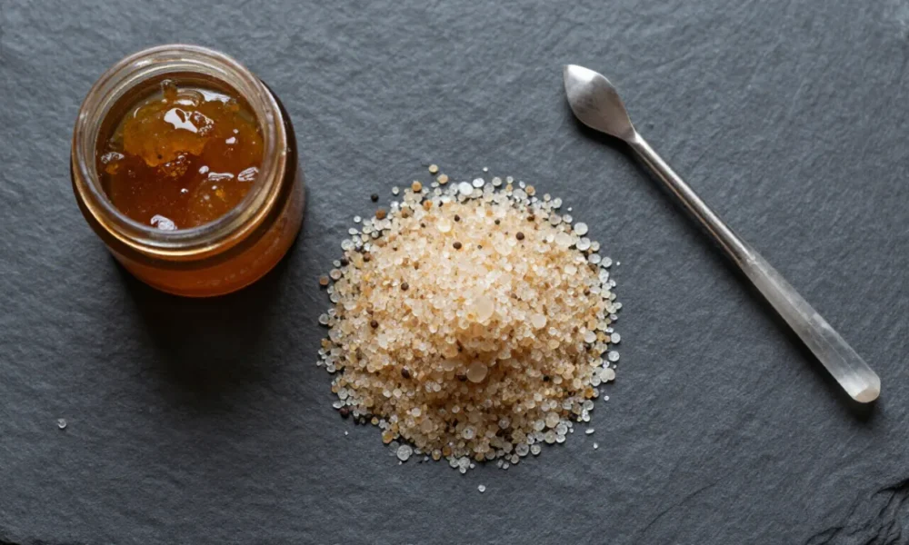 A flat lay photograph showing different types of CBD concentrates including white isolate powder, amber glass-like shatter in a jar, golden buttery wax on parchment paper, and light brown crumbly crumble, arranged on a dark slate surface with a dab tool.