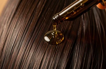 Close-up of pure batana oil being dropped onto healthy long dark hair from a glass bottle, showing its golden texture for hair nourishment and moisturizing benefits