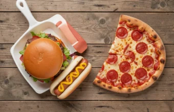 A top-down flat lay of the most popular foods in America, including a hamburger, pepperoni pizza, hot dog, mac and cheese, chocolate chip cookie, and apple pie on a rustic wooden table.