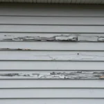 Damaged house siding with warped vinyl panels, cracks, peeling paint, and visible moisture damage on exterior wall